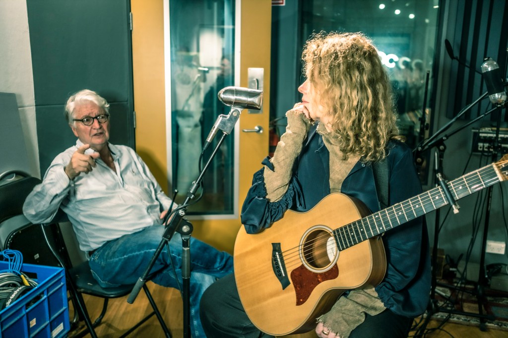 Paula and Phillip Punch in a recording studio. Paula is holding an acoustic guitar and looking back to Phillip, who is speaking and gesturing.